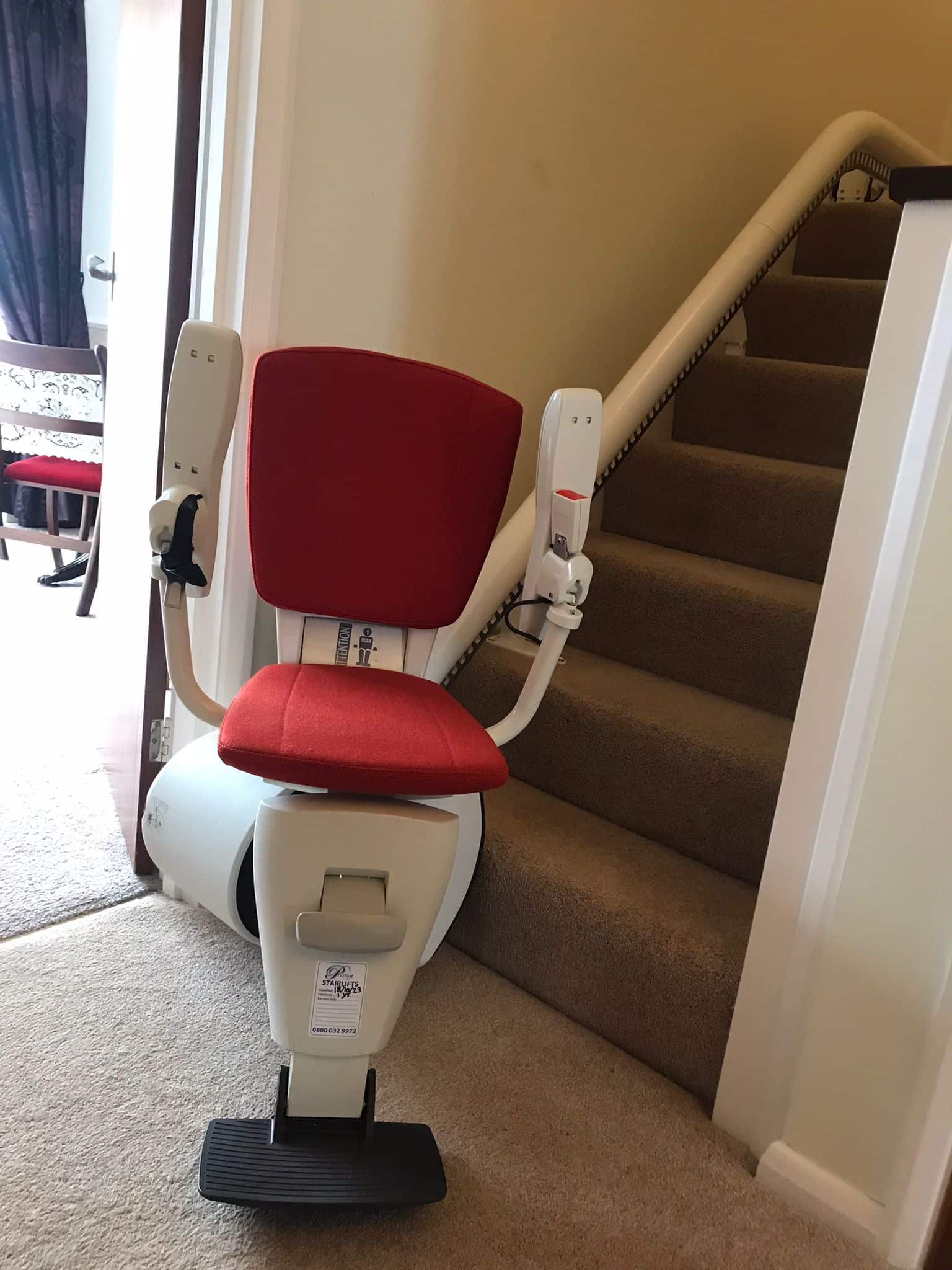 Stairlift at the bottom of stairs with red upholstered seat with arm rests in the raised position